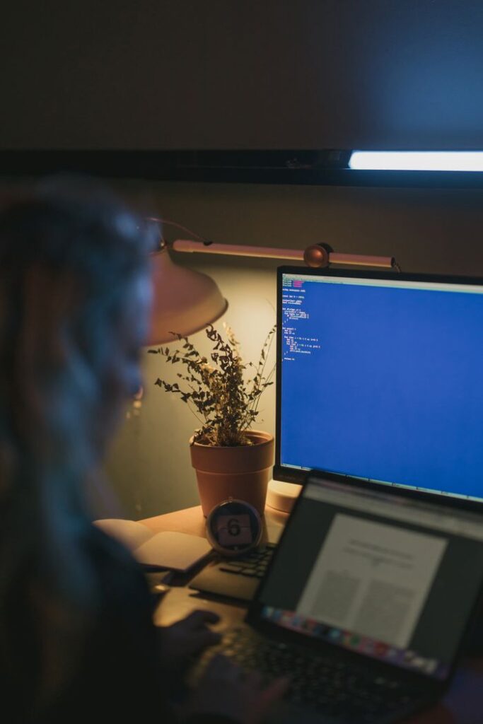 A person using a computer and laptop at night with a potted plant and desk lamp.
