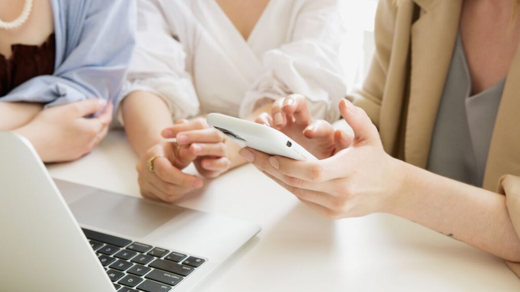 Three women collaborate using a smartphone and laptop in a professional setting.