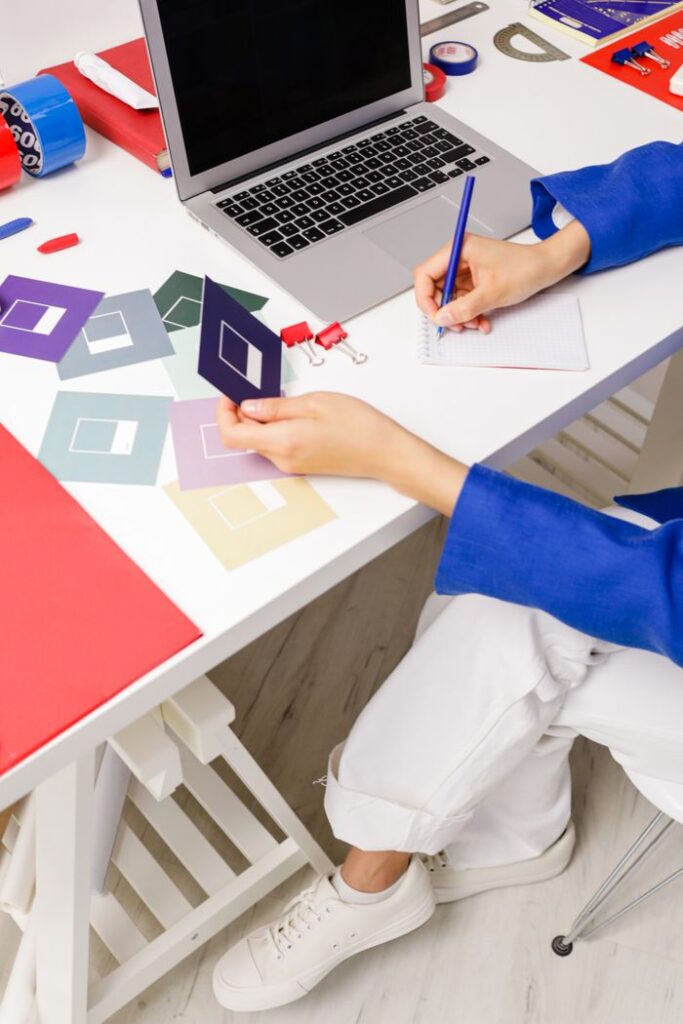 A woman writes at a desk surrounded by colorful design elements and a laptop.